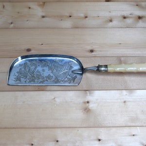 May include: A silver dustpan with an ornate design of birds and butterflies. The dustpan has a white, faux bone handle.
