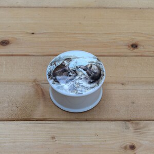 May include: A white ceramic trinket box with a round lid featuring a watercolor illustration of two otters. The lid has the word "Otters" printed on the bottom.