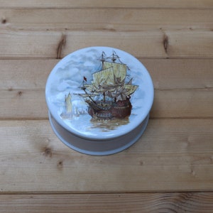 May include: A white ceramic trinket box with a round lid featuring a hand-painted image of a sailing ship with brown sails and a blue and white sky.