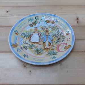 May include: A yellow and blue ceramic plate with a Peter Rabbit theme. The plate features Peter Rabbit and his sister, a garden scene, and the letters A, B, and C. The plate has the text "Which fruit begins with a?" and "Which vegetable begins with e?"