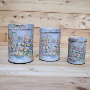 May include: Set of three round metal canisters with a white background and a colourful illustration of teddy bears sitting around a table. The canisters are different sizes.