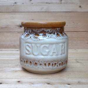 May include: A ceramic sugar jar with a wooden lid. The jar is off-white with brown accents and the word "SUGAR" embossed on the front. The jar is cylindrical with a rounded base and a wooden lid.
