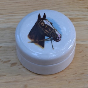 May include: A white ceramic trinket box with a round lid. The lid features a printed image of a brown horse with a bridle, set against a light blue background.