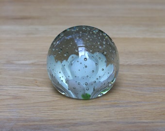 May include: A clear glass paperweight with a white flower design inside. The flower has a light pink center and is surrounded by small air bubbles.