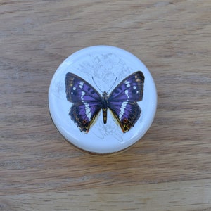 May include: A white round trinket box featuring a purple butterfly design. The butterfly has brown and white markings on its wings.