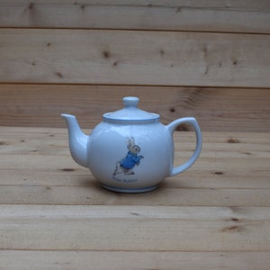May include: White ceramic teapot featuring a blue and white Peter Rabbit design. The teapot has a rounded body and a handle on the right side.