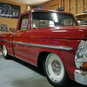 May include: A red 1967 Ford F-100 pickup truck with chrome accents and whitewall tires. The truck is parked in a garage with a black and white silhouette of a pickup truck on the wall behind it.
