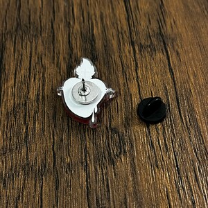 May include: A red and white enamel pin featuring a cartoon heart design. The pin has a silver metal backing and a safety clasp.