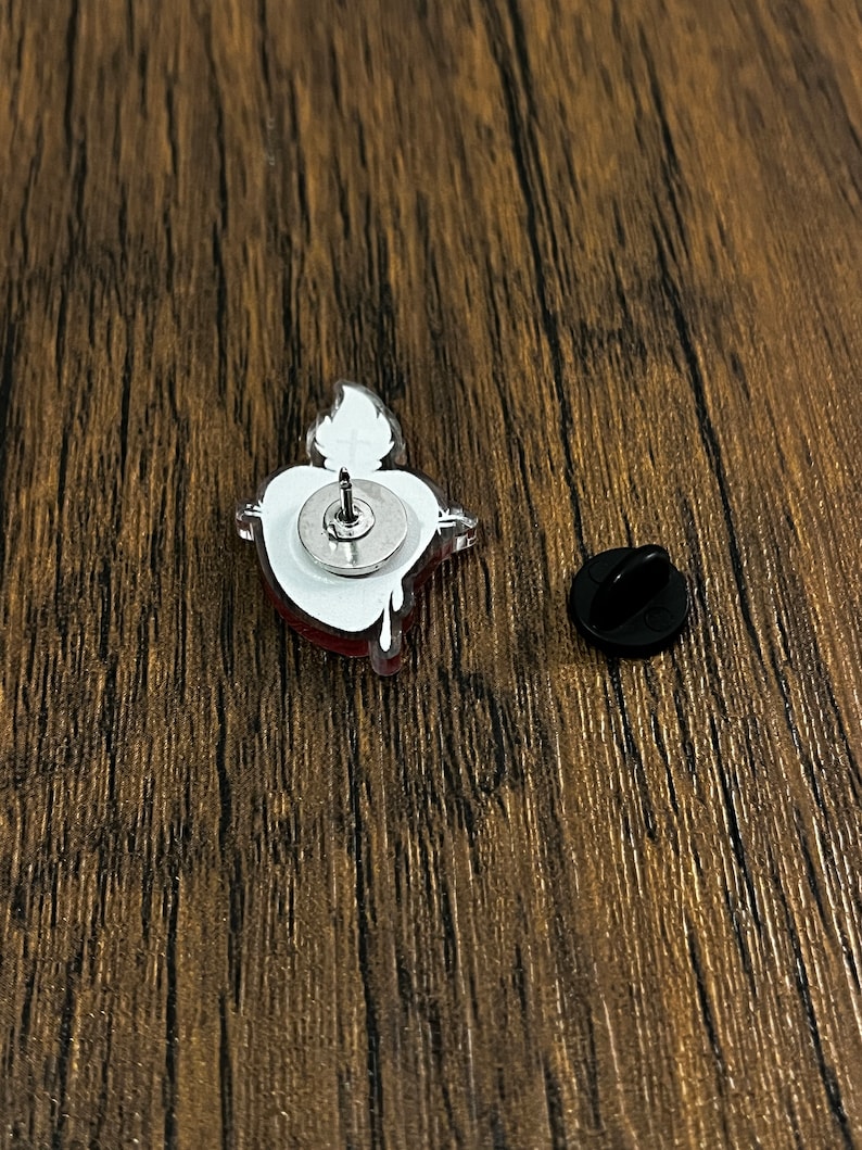 May include: A red and white enamel pin featuring a cartoon heart design. The pin has a silver metal backing and a safety clasp.