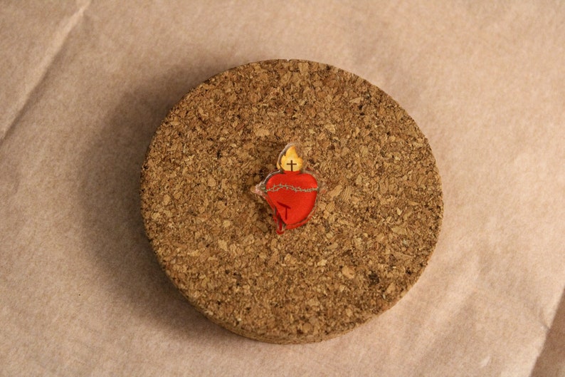 May include: A red heart with a yellow flame and a cross on top, surrounded by a brown string, on a cork background.