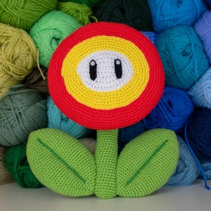 Puede incluir: Una flor de ganchillo hecha a mano al estilo de un personaje de videojuego. La flor tiene un anillo exterior rojo, un anillo central amarillo y un centro blanco con ojos ovalados negros. El tallo verde tiene dos hojas verdes. El fondo es una colección de hilos coloridos.