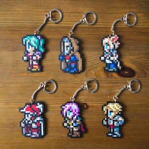 Peut inclure: Six porte-clés représentant des personnages en pixel art sur un fond en bois. Les personnages sont une femme aux cheveux verts, un homme avec une cape bleue, un homme avec une chemise bleue, une femme avec un chapeau rouge, une femme avec des cheveux violets et un homme avec des cheveux blonds.