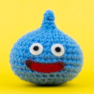 Könnte beinhalten: Ein blaues Häkel-Plüschtier in Tropfenform mit einer spitzen Oberseite. Es hat zwei runde weiße Augen mit schwarzen Pupillen und einen breiten, horizontalen roten Mund. Die Textur besteht aus sichtbaren Garnmaschen, was eine weiche Form ergibt.