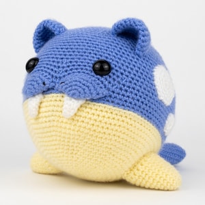 May include: A round, crocheted plush toy shaped like a walrus with a blue upper body and a pale yellow underbelly. It features two small white tusks, two black plastic eyes, small rounded ears, and white circular spots on its blue back.