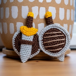 May include: Two crocheted amigurumi toys, one shaped like a sword with a yellow pommel and the other shaped like a shield with brown stripes.