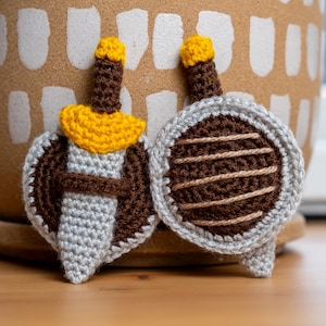 May include: Two crocheted appliques featuring a sword and a shield. The sword has a brown handle, a yellow guard, and a light gray blade. The circular shield is brown with a light gray border and features horizontal tan yarn detailing.