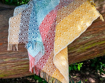May include: A woven throw blanket featuring a repeating diamond pattern in a gradient of colours including brown, blue, red, and yellow. The blanket has a textured surface and is finished with fringe along the edges, displaying a vibrant colour transition.