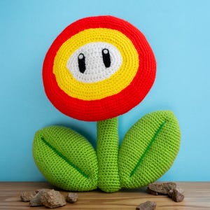 May include: A crocheted plush toy shaped like a flower with a large circular head featuring concentric rings of red, yellow, and white. The white centre has two black oval eyes. It has a green stem and two green leaves with stitched veins.
