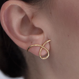 May include: Gold-toned ear cuff with a simple, abstract design. The cuff is made of a single piece of metal that is bent into a curved shape.