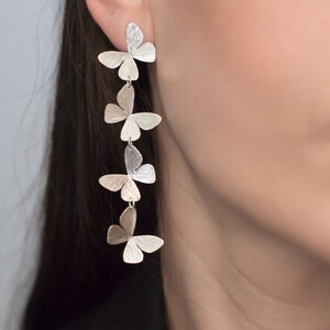 May include: Silver butterfly earrings with five butterflies dangling from each other. The butterflies are all the same size and shape, and they are arranged in a vertical line.