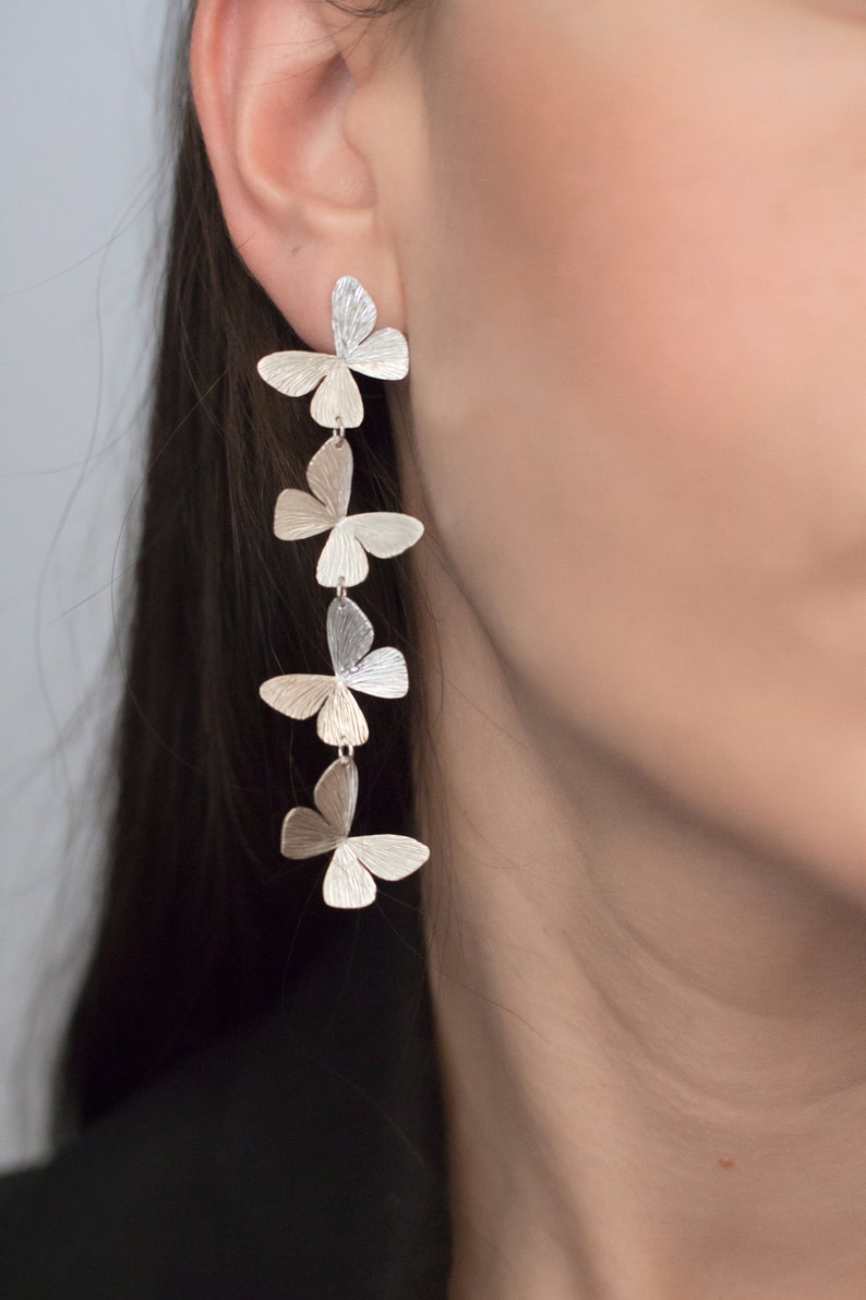 May include: Silver butterfly earrings with five butterflies dangling from each other. The butterflies are all the same size and shape, and they are arranged in a vertical line.