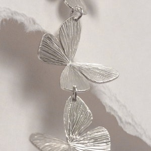 May include: Silver dangle earrings with three layered butterfly charms. Each butterfly charm has a textured surface.