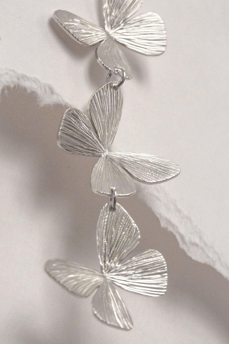 May include: Silver dangle earrings with three layered butterfly charms. Each butterfly charm has a textured surface.