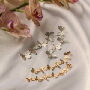 May include: Two pairs of butterfly earrings, one pair in gold and one pair in silver. The earrings are arranged on a white fabric background.