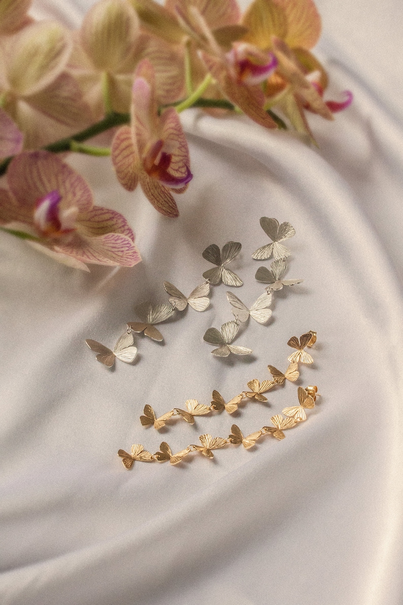 May include: Two pairs of butterfly earrings, one pair in gold and one pair in silver. The earrings are arranged on a white fabric background.