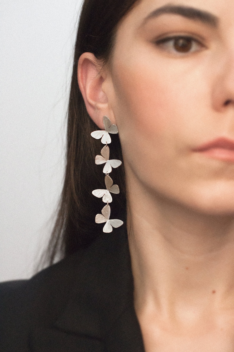 May include: Silver butterfly earrings with a layered design. The earrings are long and dangle from the earlobe.