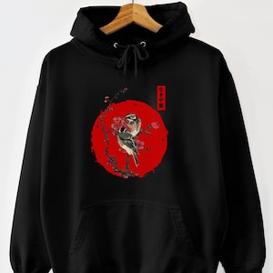 Op de afbeelding: Zwarte hoodie met een Japans geïnspireerd ontwerp. Een grote rode cirkel met een vogel op een bloeiende tak is gecentreerd op de voorkant. De hoodie heeft een capuchon met trekkoord en een voorzak.