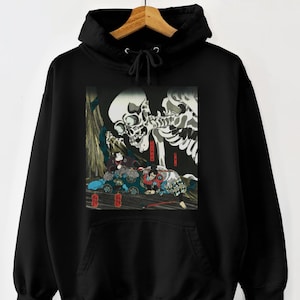 Op de afbeelding: Zwarte hoodie met een grafische print met een skeletfiguur en figuren in traditionele Japanse kleding. Het kunstwerk is in vintage stijl, op een houten hanger.