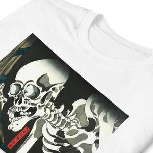 May include: White t-shirt featuring a detailed illustration of a skeleton in a traditional Japanese art style. The artwork is predominantly black, white, and gray, with a red rectangular text box. The shirt has a classic crew neck.