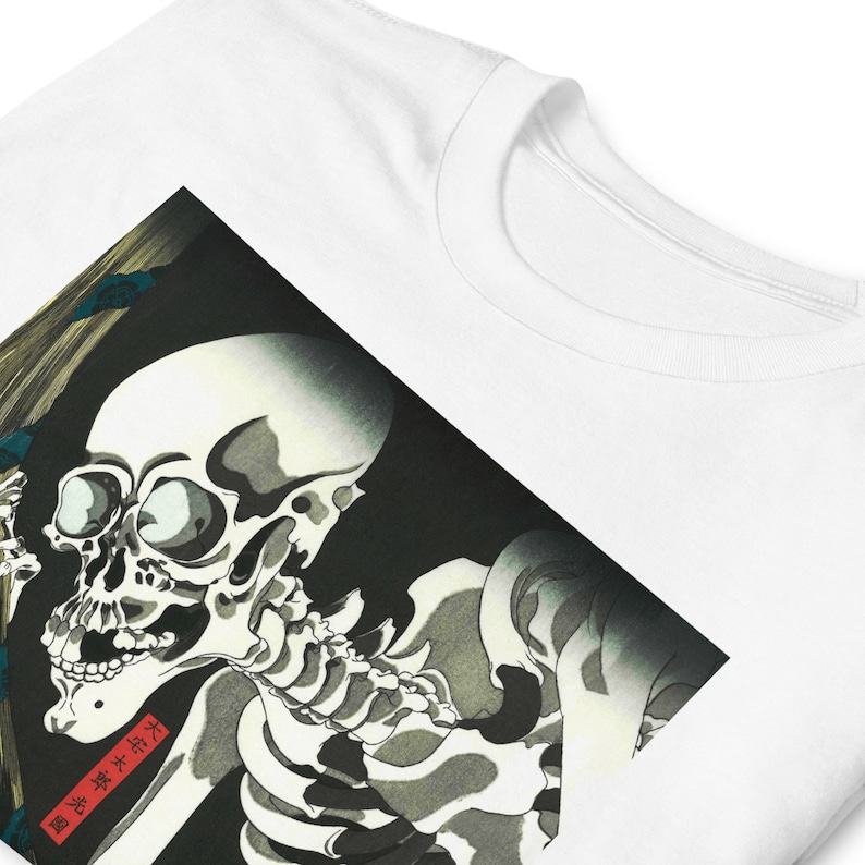 May include: White t-shirt featuring a detailed illustration of a skeleton in a traditional Japanese art style. The artwork is predominantly black, white, and gray, with a red rectangular text box. The shirt has a classic crew neck.
