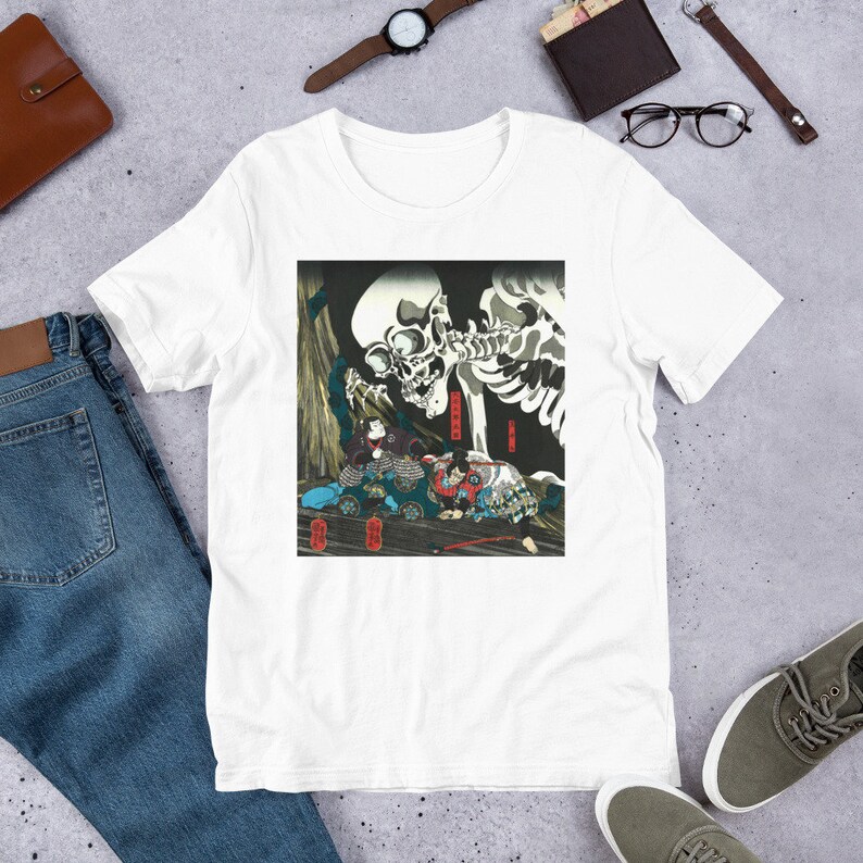 May include: White t-shirt featuring a Japanese woodblock print of a skeletal figure looming over several figures in traditional clothing. The artwork is in shades of black, red, and blue. The shirt is made of a soft material.