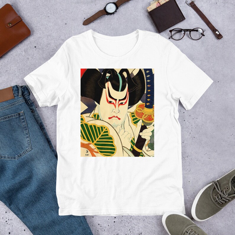 以下が含まれることがあります： 剣を持った日本の侍のカラフルな画像がプリントされた白いTシャツ。侍は険しい表情で、伝統的な服装をしています。