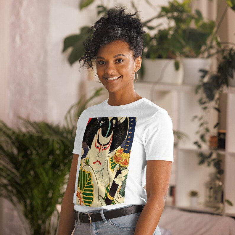 以下が含まれることがあります： 剣を持った侍のカラフルな日本の木版画がプリントされた白いTシャツ。プリントは赤、青、緑、金色の詳細なデザインです。