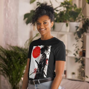 May include: Black t-shirt featuring a graphic design of a samurai warrior with a red sun and a sword. The artwork is in black, white, and red. The t-shirt is made of a soft material.