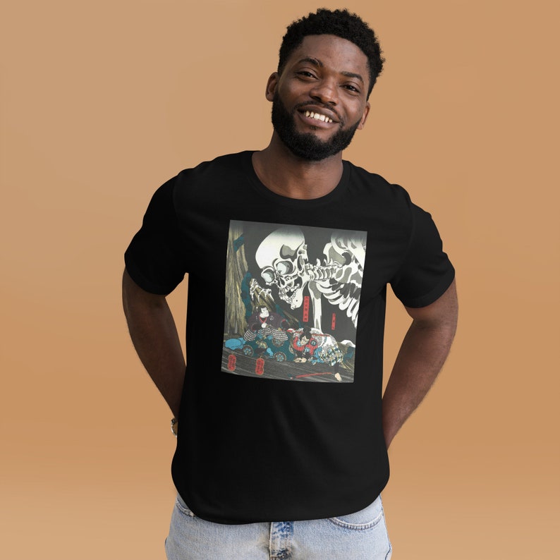 May include: Black t-shirt featuring a graphic print of a skeleton and figures in a traditional Japanese art style. The artwork includes a large skull and figures in colorful clothing. The t-shirt is made of a soft material.