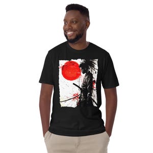 May include: Black t-shirt featuring a graphic of a samurai warrior with a sword, a red sun, and red splatter effects. The design is on a white background. The shirt is short-sleeved.