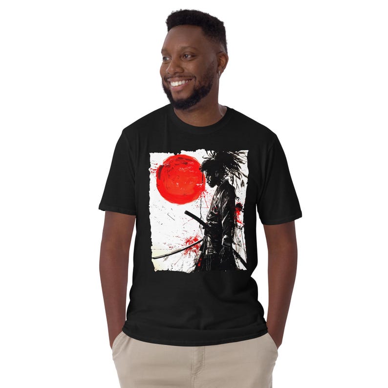 May include: Black t-shirt featuring a graphic of a samurai warrior with a sword, a red sun, and red splatter effects. The design is on a white background. The shirt is short-sleeved.
