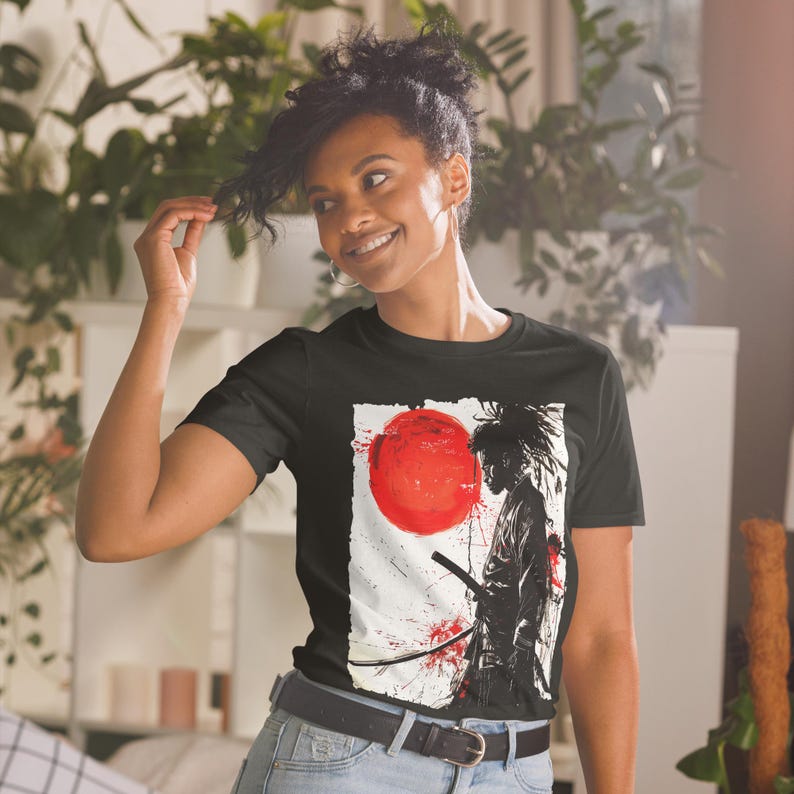 May include: Black t-shirt featuring a graphic of a samurai warrior with a sword, a red sun, and blood splatter design. The artwork is set against a white background. The shirt is worn by a person.