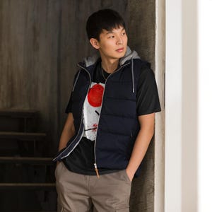 May include: A navy blue hooded puffer vest with a gray lining is worn over a black t-shirt. The t-shirt features a graphic design with a large red circle and white and black accents. The person is wearing brown cargo pants.