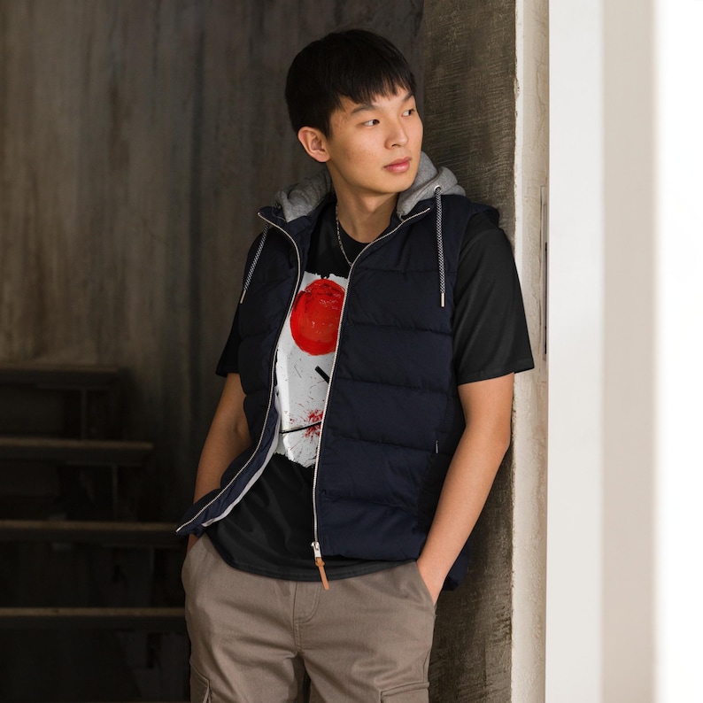 May include: A navy blue hooded puffer vest with a gray lining is worn over a black t-shirt. The t-shirt features a graphic design with a large red circle and white and black accents. The person is wearing brown cargo pants.