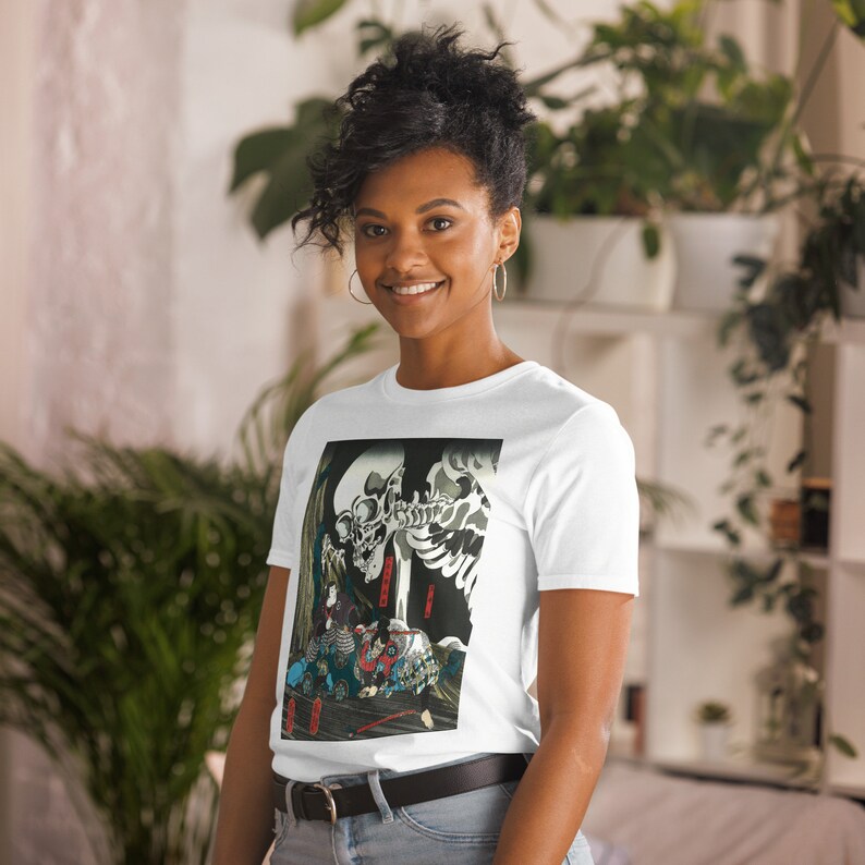 May include: White t-shirt featuring a Japanese-style illustration of a skeleton and figures in traditional clothing. The artwork is predominantly black, white, and red, with accents of blue and green. The shirt is a short-sleeve style.