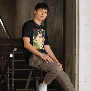 以下が含まれることがあります： 戦士の顔のカラフルな日本のスタイルのプリントが施された黒いTシャツ。シャツを着ている人は、スツールに座っています。