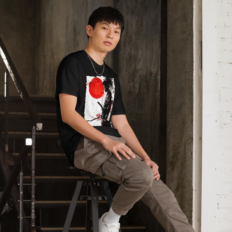 May include: Black t-shirt featuring a Japanese-inspired graphic with a red sun and a samurai figure. The design is set against a white background with black and red accents. The shirt is made of a soft material.