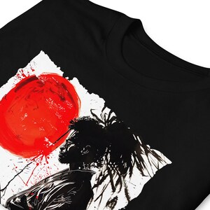 May include: Black t-shirt featuring a striking graphic design. The design includes a large red circle, a black silhouette of a person, and white and red accents. The artwork has a bold, artistic style.