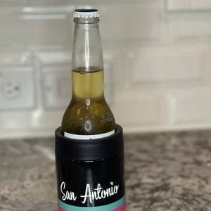K&ouml;nnte beinhalten: Ein schwarzer und rostfreier Dosenk&uuml;hler mit einem wei&szlig;en Design, auf dem "San Antonio" mit drei horizontalen Streifen in Orange, Pink und T&uuml;rkis steht. Eine Bierflasche befindet sich im K&uuml;hler.