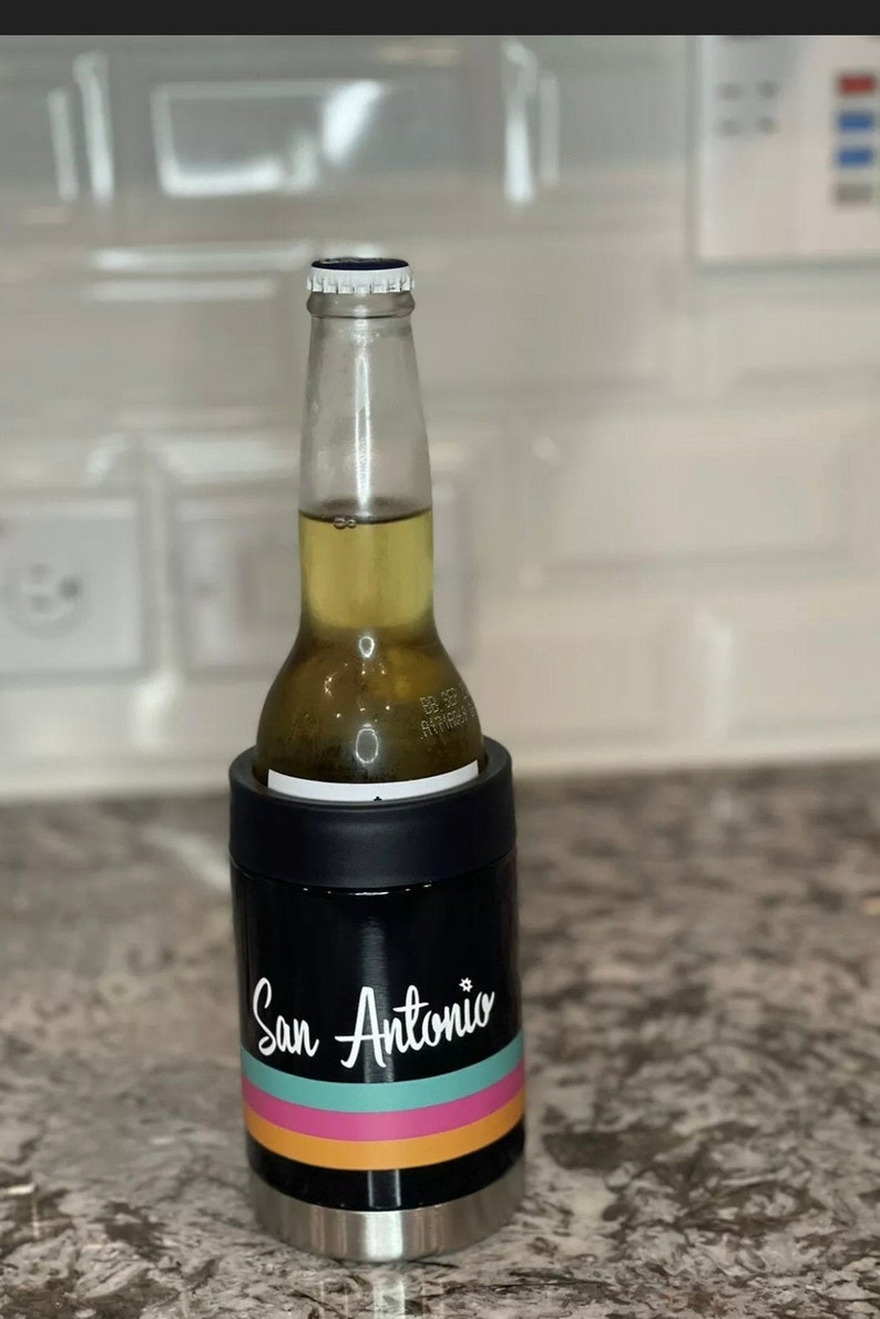 K&ouml;nnte beinhalten: Ein schwarzer und rostfreier Dosenk&uuml;hler mit einem wei&szlig;en Design, auf dem "San Antonio" mit drei horizontalen Streifen in Orange, Pink und T&uuml;rkis steht. Eine Bierflasche befindet sich im K&uuml;hler.